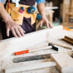 Hombre carpintero trabajando en un taller, midiendo y ajustando una pieza de madera con herramientas de carpintería como un martillo y un nivel, rodeado de virutas de madera.