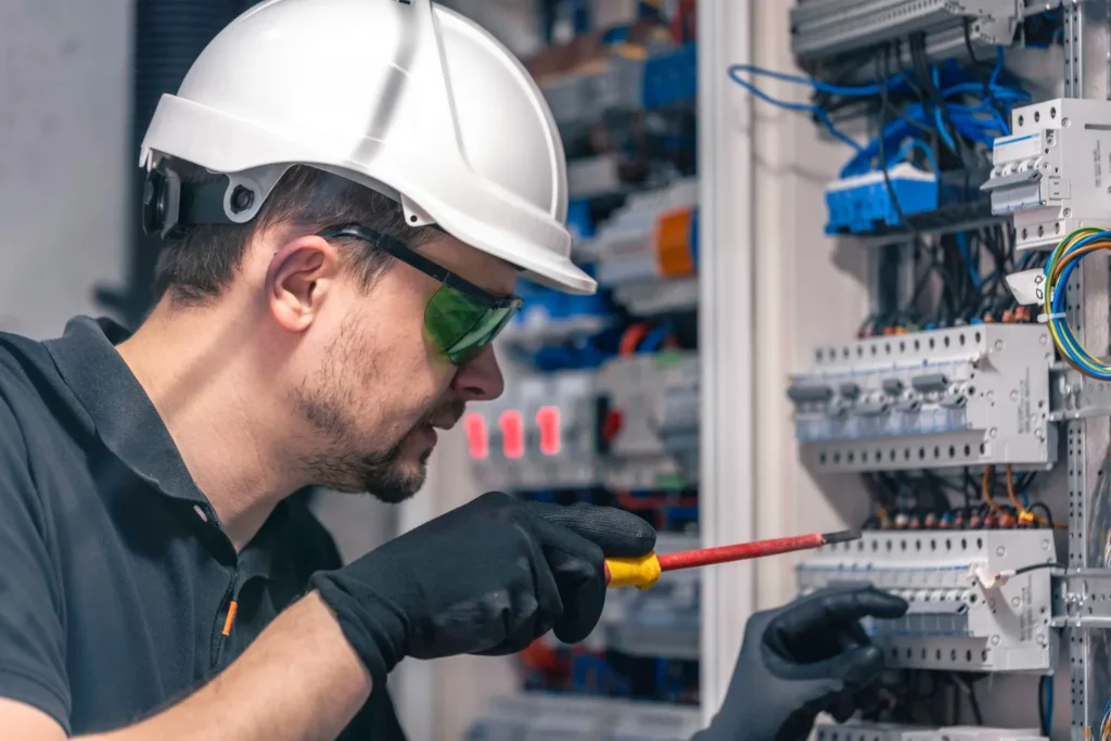 Técnico electricista trabajando en un panel eléctrico, usando gafas de seguridad y casco. Se observa el uso de un destornillador para realizar conexiones eléctricas.
