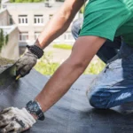 Trabajador en el techo instalando una membrana impermeable. Se observan manos con guantes sosteniendo el material mientras se aplica sobre la superficie. Fondo de edificios y vegetación.